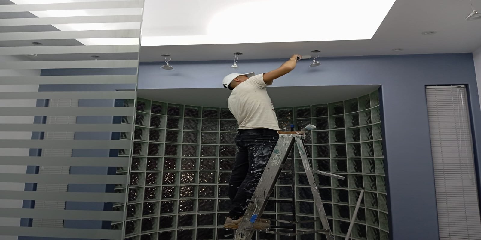 Hombre pintando techo sobre escalera en oficina moderna.