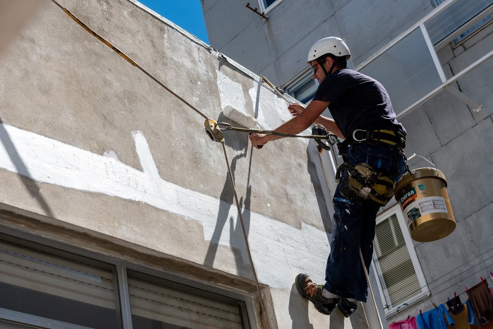 Trabajador pintando fachada, colgado con arneses.